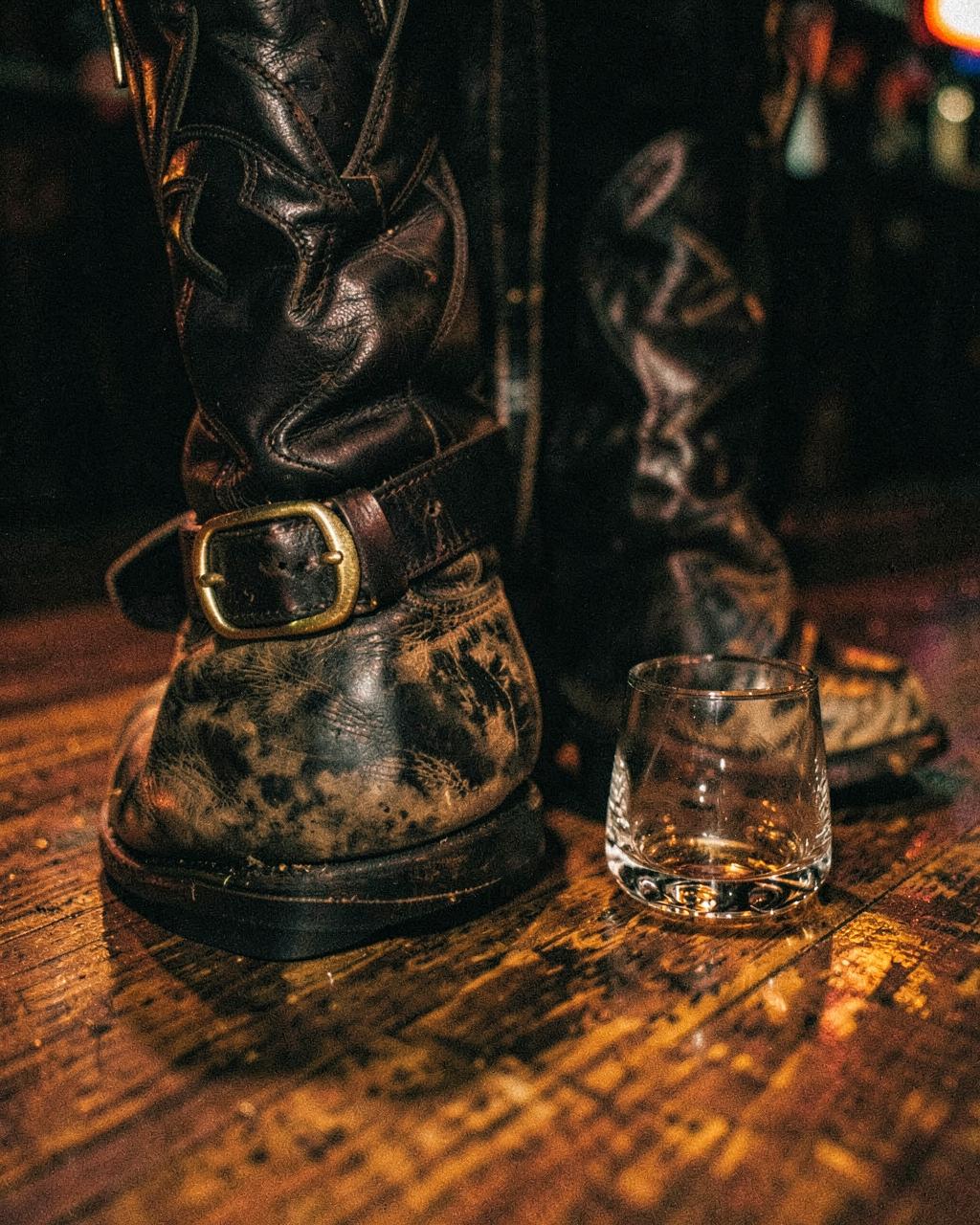 Close-up of cowboy boots, a gold belt buckle and a whiskey glass